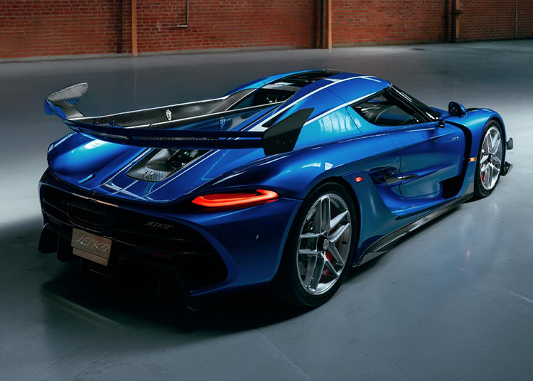 A blue Koenigsegg Jesko hypercar with a large rear spoiler is parked in an industrial-style garage with brick walls and a concrete floor.