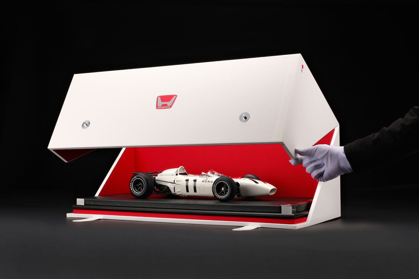 A gloved hand opens a white display case with a red interior, revealing a detailed model of the RA272 inside—a tribute to the legacy and honor of vintage race cars.