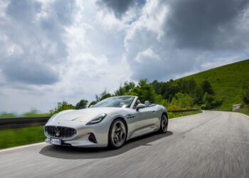 A white convertible sports car drives on a winding road through a green, hilly landscape under a cloudy sky.