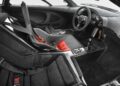 Interior of a sports car with a racing seat, seat belts, exposed carbon fiber, manual gear shifter, and a detailed dashboard with analog gauges—much like those found in 10 Cars That Feature a Central Driving Position.