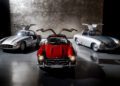 Three classic Mercedes-Benz cars with gullwing doors open, including the iconic 300 SL—the Blue Chip Benz—are displayed in a dimly lit showroom with a textured wall background.