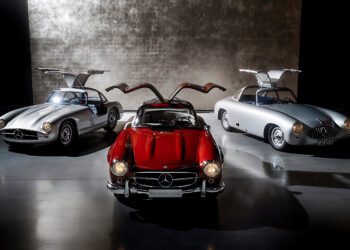 Three classic Mercedes-Benz cars with gullwing doors open, including the iconic 300 SL—the Blue Chip Benz—are displayed in a dimly lit showroom with a textured wall background.