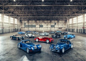 A group of classic-style sports cars, including the Morgan 25th Anniversary edition, mostly blue with a few red and silver, parked inside a spacious industrial warehouse with high ceilings and large windows.