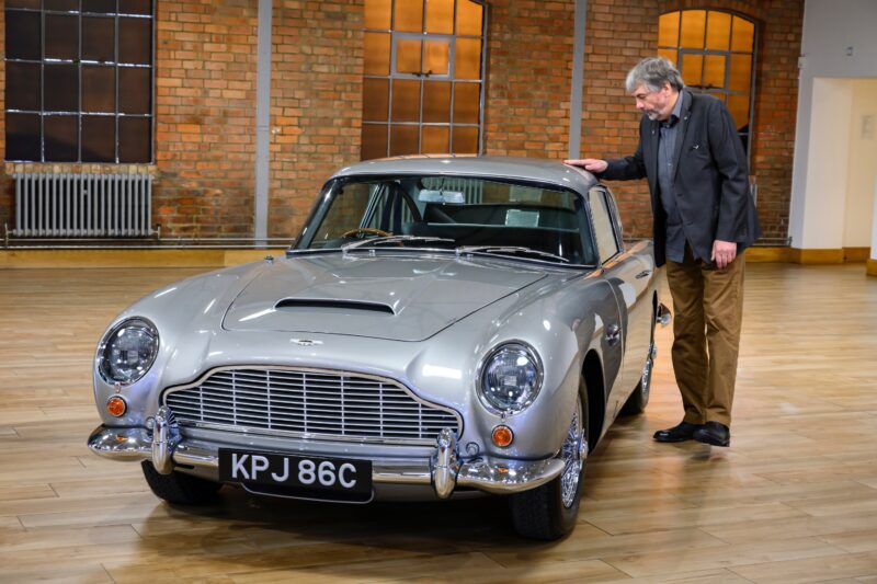 A man stands next to a classic silver Aston Martin DB5—revived by Aston Martin Works after a 50 year wait—its KPJ 86C license plate hinting at an incredible story, all set in a spacious room with brick walls and large windows.
