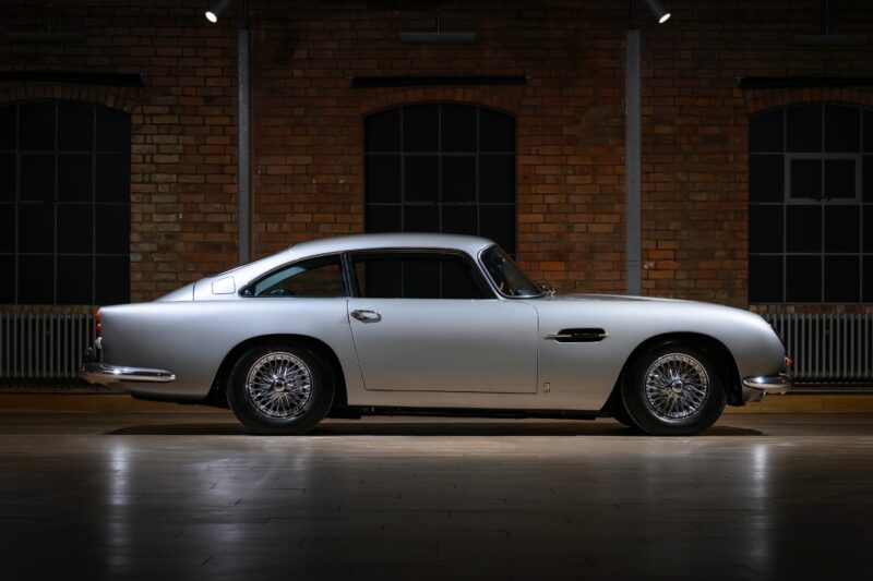 Side view of a rare Aston Martin DB5 coupe in silver, parked indoors against a brick wall with large windows—a stunning example of classic design from Aston Martin Works.