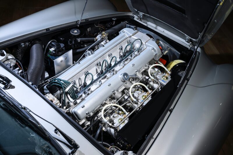 A close-up view of a classic Aston Martin DB5’s engine bay, showcasing an inline-six engine with polished metal components and visible fuel lines, surrounded by a silver-painted chassis.