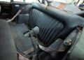 The image shows the worn and dirty backseat of a rare old car, possibly an Aston Martin Works DB5, with torn upholstery, exposed seatbelts, and visible signs of age and neglect.