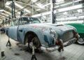 A rusted, partially disassembled rare DB5 is lifted on jacks inside a well-lit auto repair workshop, with other vehicles in the background, hinting at an incredible story reminiscent of Aston Martin Works.