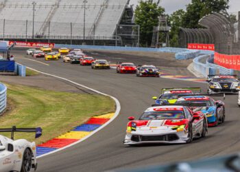 A group of race cars, including the Ferrari of Seattle, speed around a curved section of a racetrack, with empty grandstands and green trees in the background during the Ferrari Challenge.