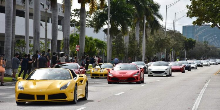 A line of luxury sports cars, including several Ferraris, drives down a palm-lined city street with people gathered on the sidewalk.