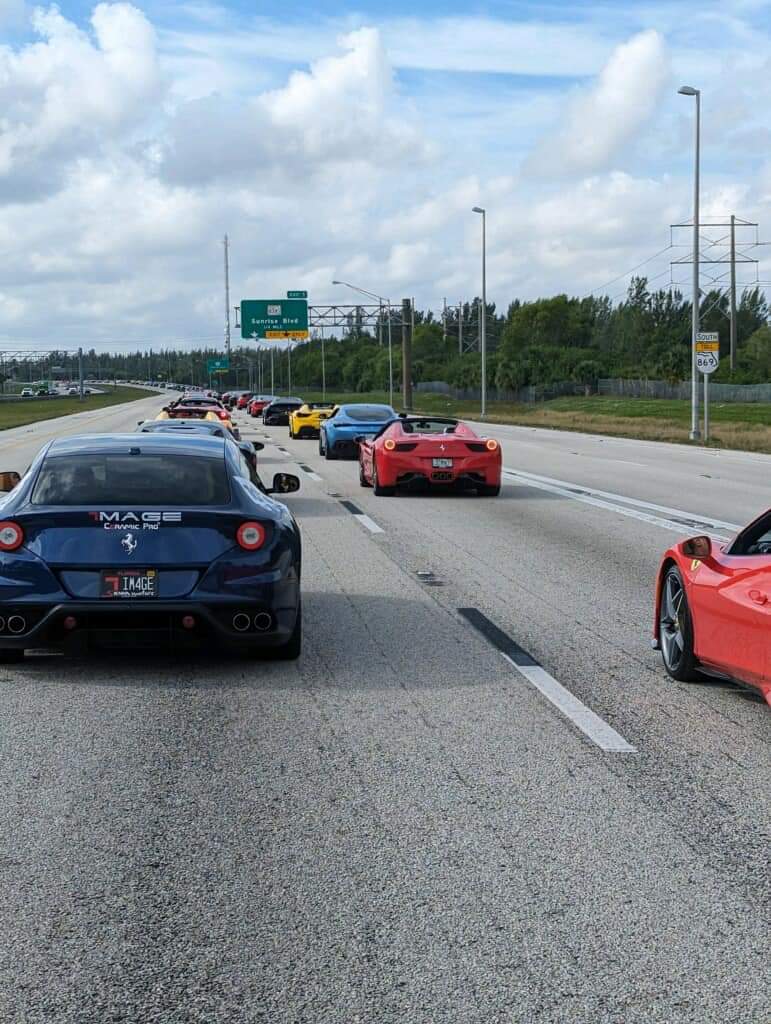 A group of luxury sports cars, including several Ferraris, drives together on a multi-lane highway under a partly cloudy sky.