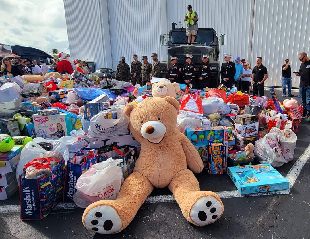 Large pile of donated toys, including a giant teddy bear, with uniformed military personnel and volunteers standing nearby outdoors.