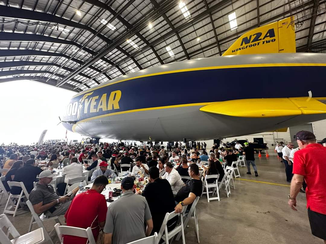 People sit at tables under the Goodyear blimp inside a large hangar during an indoor event.