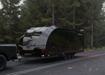 A black Race Transporter 7 enclosed car hauler labeled "RT17" is being towed by a dark pickup truck on a forested road, showcasing the quality of Brian James Trailers.