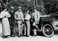 Five people stand beside an early 20th-century Rolls-Royce on a road, dressed in period clothing, with trees in the background, as Rolls-Royce marks 150 years of Charles Sykes, the artist behind the Spirit of Ecstasy.