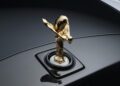 Close-up of a gold Spirit of Ecstasy hood ornament on the glossy black hood of a Rolls-Royce, celebrating 150 years of Charles Sykes, the artist behind this iconic symbol.