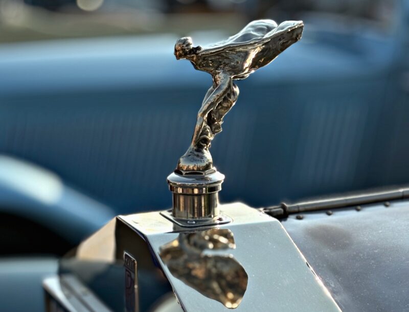 Close-up of a chrome hood ornament shaped like a woman leaning forward, mounted on a vintage car—celebrating Rolls-Royce Marks 150 Years of Charles Sykes, the artist behind the Spirit of Ecstasy—with a blue vehicle in the background.