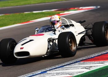 A classic white RA272 open-wheel race car with the number 11, driven by a helmeted driver, takes a turn on a racetrack with colorful curbs, honoring its legacy in motorsport history.