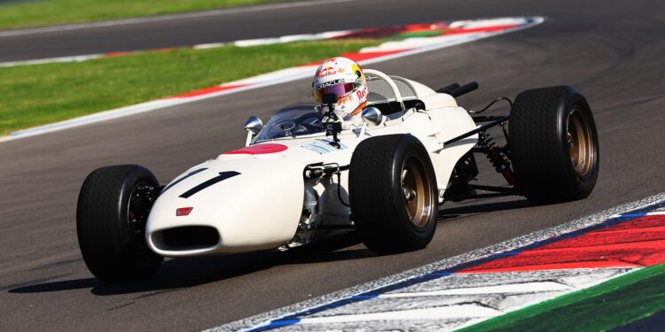 A classic white RA272 open-wheel race car with the number 11, driven by a helmeted driver, takes a turn on a racetrack with colorful curbs, honoring its legacy in motorsport history.