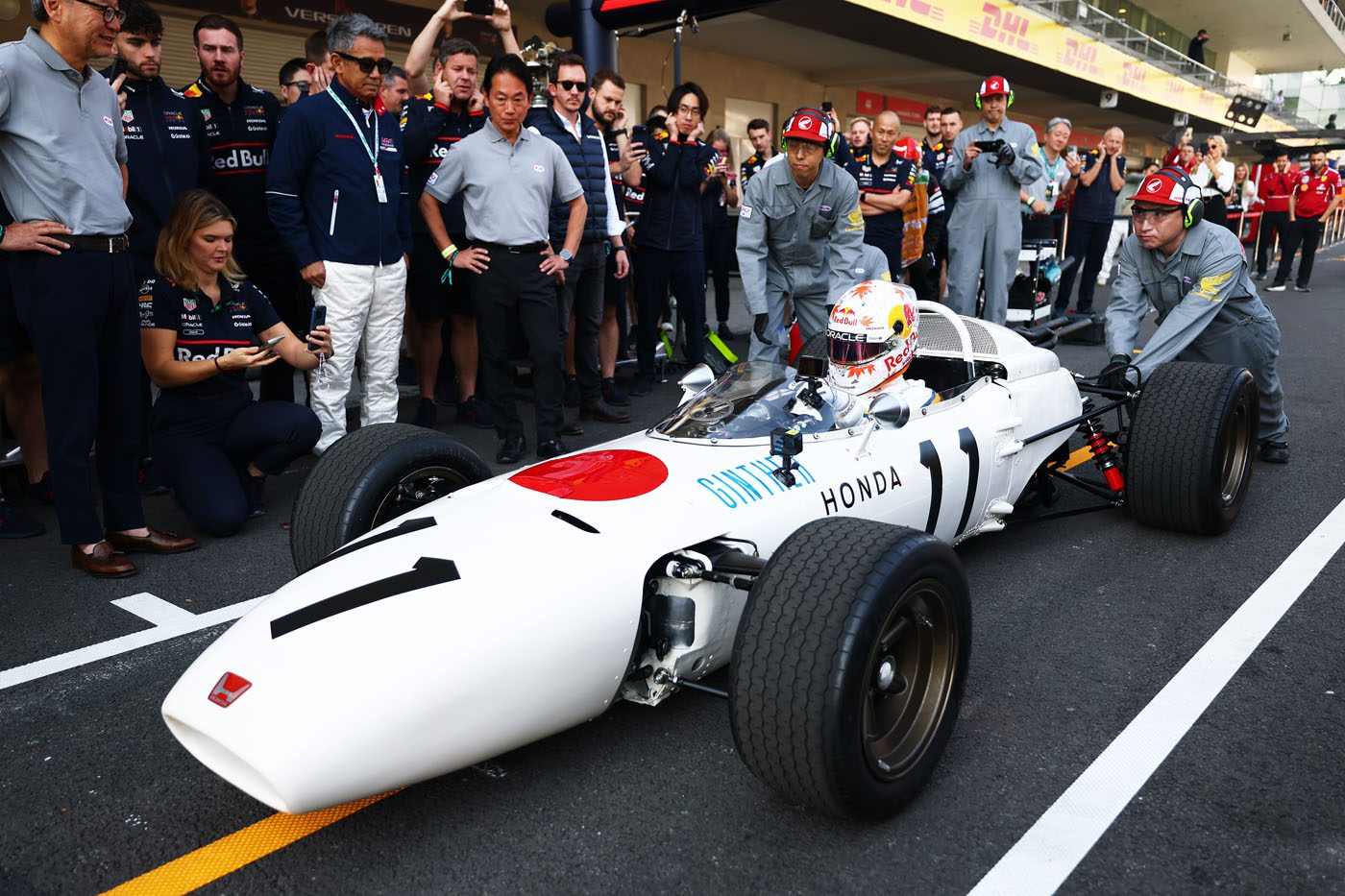 A classic white Honda RA272 Formula 1 car with a red circle is pushed by crew members in the pit lane, surrounded by people—an enduring symbol of racing honor and legacy.