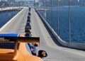 A line of sports cars, led by an orange car, drives across a long bridge over water toward a city in the distance during the Dimmitt Automotive Group Exotic Car Rally.