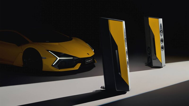 A yellow sports car is parked next to two tall, yellow and black speakers on a dark background with a stripe of light—a scene worthy of the duPont REGISTRY Holiday Gift Guide for 2026.