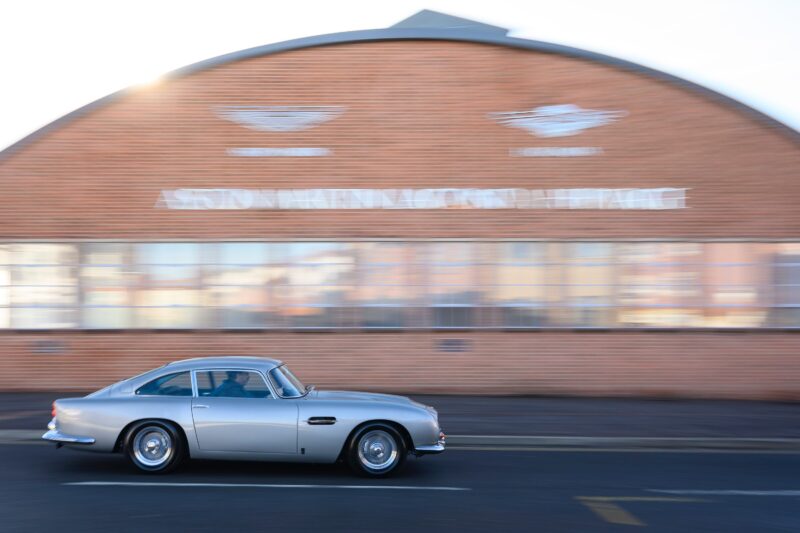 A rare DB5 silver classic car drives past a red brick building with "Aston Martin" signage in the background, capturing an incredible story at Aston Martin Works.