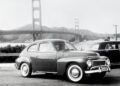 A classic car is parked near the Golden Gate Bridge on a cloudy day, evoking nostalgia for the wagon era, with another car and hills visible in the background.