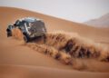 A four-wheel drive SUV drives up a sand dune in a desert, kicking up a large spray of sand behind its tires.
