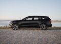 A black Volvo V60 Cross Country wagon is parked on a gravel road next to a body of water, with a clear sky in the background.