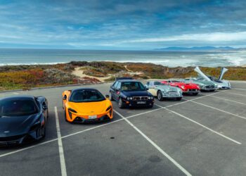 Six cars, including classic and modern models, are parked in a row near a beach with ocean waves and a cloudy sky in the background.