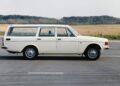 A white Volvo V60 Cross Country station wagon is parked on a paved road, evoking the wagon era amid grass fields and trees in the background.