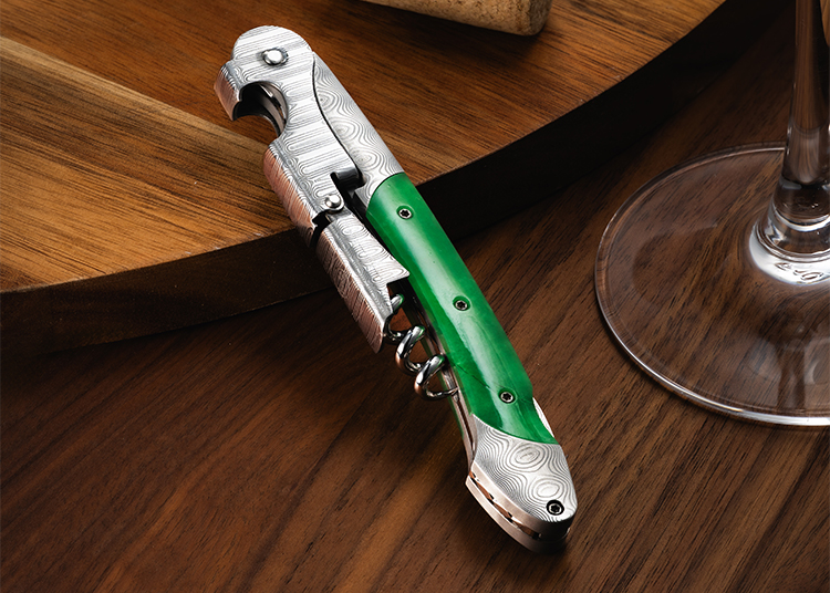 A metal corkscrew with a green handle rests on a wooden surface next to a wine glass—a perfect addition to any Holiday Gift Guide, as featured in duPont REGISTRY's 2026 recommendations.