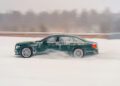A dark green luxury sedan driving quickly through snow, creating a cloud of snow behind it, with a blurred winter landscape in the background.