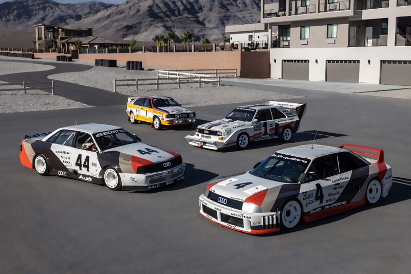 Four retro-inspired Audi race cars, including an RS3, with vintage liveries are parked in an outdoor area near modern buildings and mountains under clear daylight.