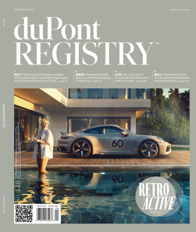 A woman in a white robe stands by a pool using her phone; a Porsche sports car with the number 60—a nod to duPont REGISTRY Cover Cars—waits beside her; a modern house completes the luxurious scene.