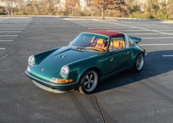 A green vintage Porsche 911 Targa, customized by Singer with a red roof and tan interior, is parked in an empty lot—a true duPont REGISTRY classic.