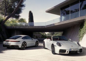 Two silver Porsche 911 sports cars, one coupe and one convertible, parked outside a modern house surrounded by greenery—a perfect showcase for Porsche’s 2025 reveals.