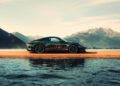 A black Porsche 911 GT3 with the number 72 and Porsche logo on the side is parked by a body of water with mountains in the background, celebrating F. A. Porsche's 90th Birthday in style.