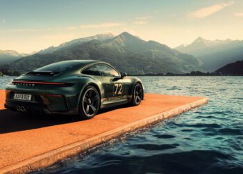 A green Porsche 911 GT3 sports car with the number 72 is parked on a concrete platform beside a lake, mountains rising behind—a striking tribute for F. A. Porsche’s 90th birthday under a partly cloudy sky.