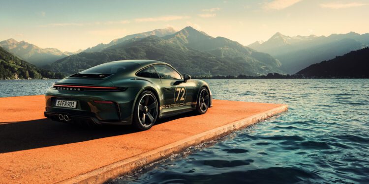 A green Porsche 911 GT3 sports car with the number 72 is parked on a concrete platform beside a lake, mountains rising behind—a striking tribute for F. A. Porsche’s 90th birthday under a partly cloudy sky.