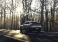 A silver Porsche Macan Electric drives on a paved road through a forest with bare trees at sunrise or sunset, capturing the magic of the Road to Christmas.