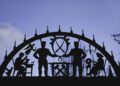 Metal archway features silhouettes of workers performing various tasks against a blue sky, with decorative lights outlining the arch—an inspiring scene on the Road to Christmas, perfect for electric car enthusiasts.