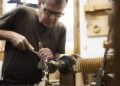 A man wearing glasses uses a chisel to shape wood on a lathe in a workshop, with wood shavings falling below—crafting each detail as meticulously as if restoring a classic Porsche for the Road to Christmas.