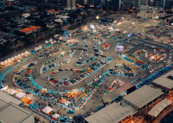 Aerial view of a large outdoor go-kart racing track surrounded by parked cars, spectators, and city buildings at dusk.