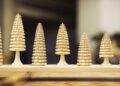 Seven handcrafted wooden trees of varying sizes are arranged in a row on a wooden surface, evoking the magic of the Road to Christmas against a blurred background.