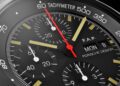 Close-up of a Porsche Design chronograph watch face, inspired by the Porsche 911 GT3, showing time, three subdials, tachymeter, and day-date display. The main hands are yellow and red.