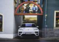 A white Porsche Macan Electric is parked beneath a decorative arch with large figurines, set between two colorful buildings on the Road to Christmas.