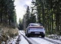 A white Porsche sedan drives along a narrow, snow-dusted forest road lined with tall evergreen trees under a cloudy sky, capturing the spirit of the Road to Christmas.