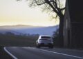 A white Porsche Macan Electric drives on a winding rural road at dusk, passing by a barn, with hills and trees in the background—a perfect scene for the road to Christmas.